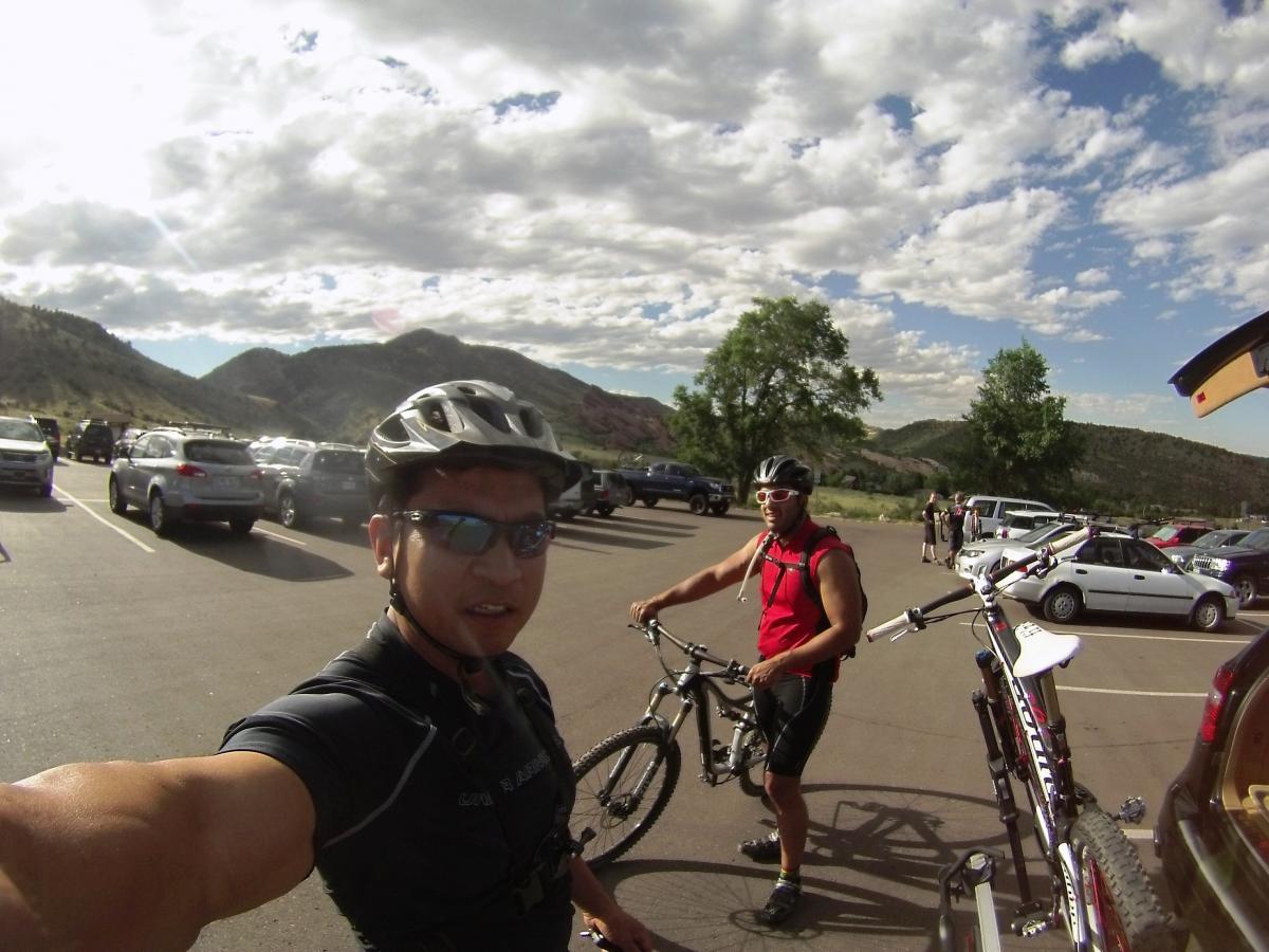 Two mountain bikers preparing to ride in a parking lot surrounded by mountains. One biker, wearing a black helmet and sunglasses, poses for a selfie while the other, in a red sleeveless top, adjusts his bike. The background features a variety of parked cars and scenic hills under a partly cloudy sky. Mount Falcon Park mountain bike trail.