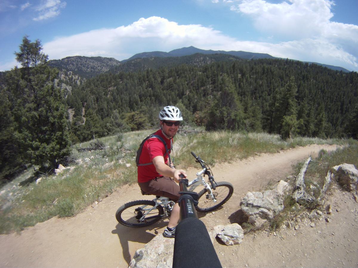 A smiling cyclist in a red shirt and helmet takes a selfie while perched on a mountain bike on a dirt trail. Lush green hills and a blue sky with fluffy clouds create a picturesque backdrop. The scene captures the spirit of outdoor adventure and mountain biking. Betasso Preserve mountain bike trail.