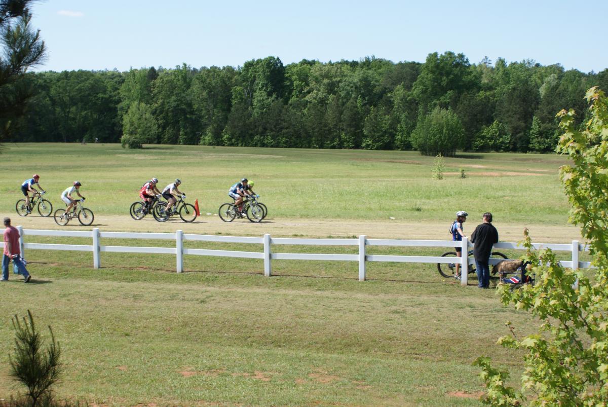 A group of six cyclists riding on a dirt path in a green landscape, with trees in the background. A man is walking nearby, and two people are standing by a white fence, one of whom appears to be talking to a cyclist. The scene is bright and sunny, showcasing a grassy area with a few scattered trees. Georgia International Horse Park mountain bike trail.