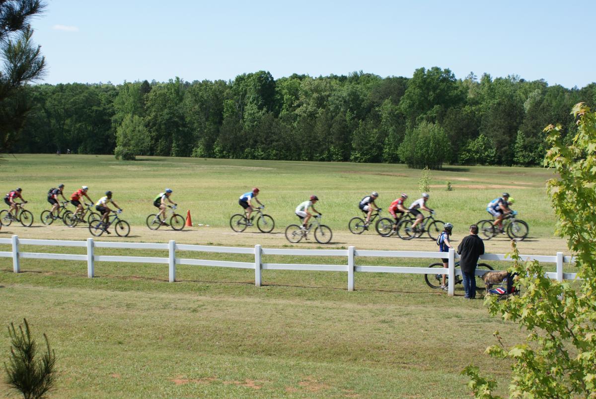 A group of cyclists riding along a dirt path in a park, surrounded by green trees and open fields. A person stands next to a bicycle by a white fence, watching the cyclists as they pass by. Bright blue skies are visible above. Georgia International Horse Park mountain bike trail.