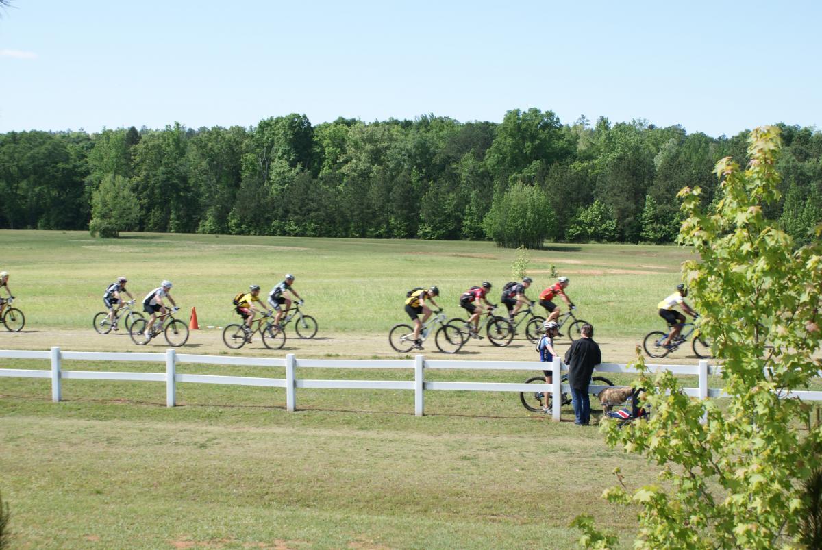 A group of cyclists riding in a mountain bike race on a dirt path, with greenery in the background and a few spectators watching from the side. A bicycle is parked nearby, and there are orange cones marking the course. Georgia International Horse Park mountain bike trail.