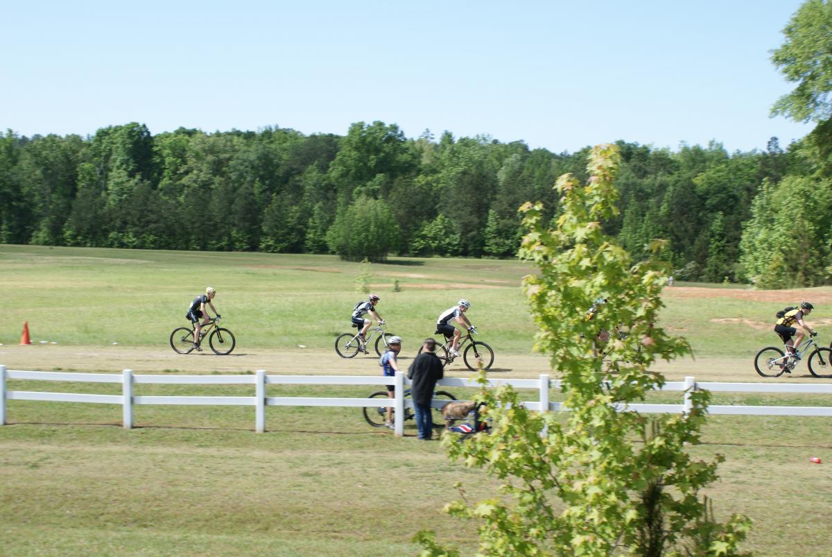 A group of cyclists riding on a dirt path in a green park area. In the foreground, a person is standing beside a bicycle while a small dog is nearby. There are trees in the background and a white fence along the path, with an orange cone visible on the ground. The sky is clear and sunny. Georgia International Horse Park mountain bike trail.