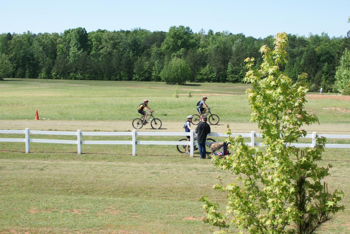 A scenic outdoor setting featuring three cyclists riding on a dirt path surrounded by greenery. In the foreground, a person stands talking to another, while a bicycle rests nearby. A white fence and a small tree are also visible, with a clear blue sky overhead. Georgia International Horse Park mountain bike trail.