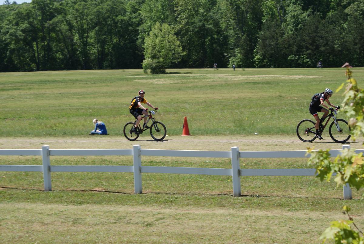 Two cyclists ride on a gravel path through a grassy area, with a white fence in the foreground. One cyclist is dressed in a black and red outfit, while the other wears a yellow and black jersey. In the background, a person sits on the ground, and additional cyclists can be seen further away among the trees. Georgia International Horse Park mountain bike trail.