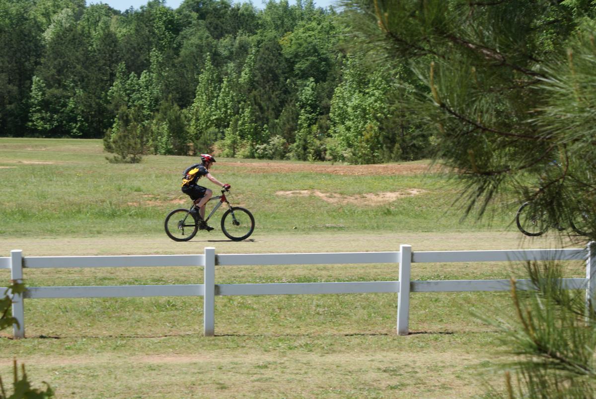 A person riding a mountain bike along a grassy path, with a white wooden fence in the foreground and a backdrop of trees. The scene is set on a sunny day, showcasing a vibrant outdoor environment. Georgia International Horse Park mountain bike trail.