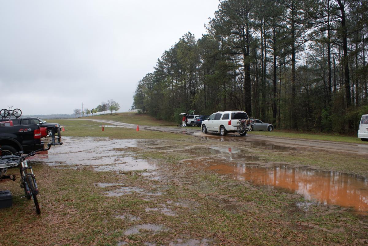 A wet and muddy parking area surrounded by trees, with several parked vehicles, including a white SUV with bicycles mounted on a rack. Rain has created puddles on the ground, and the sky is overcast. Some orange traffic cones are visible along the side of the road. Heritage Park mountain bike trail.