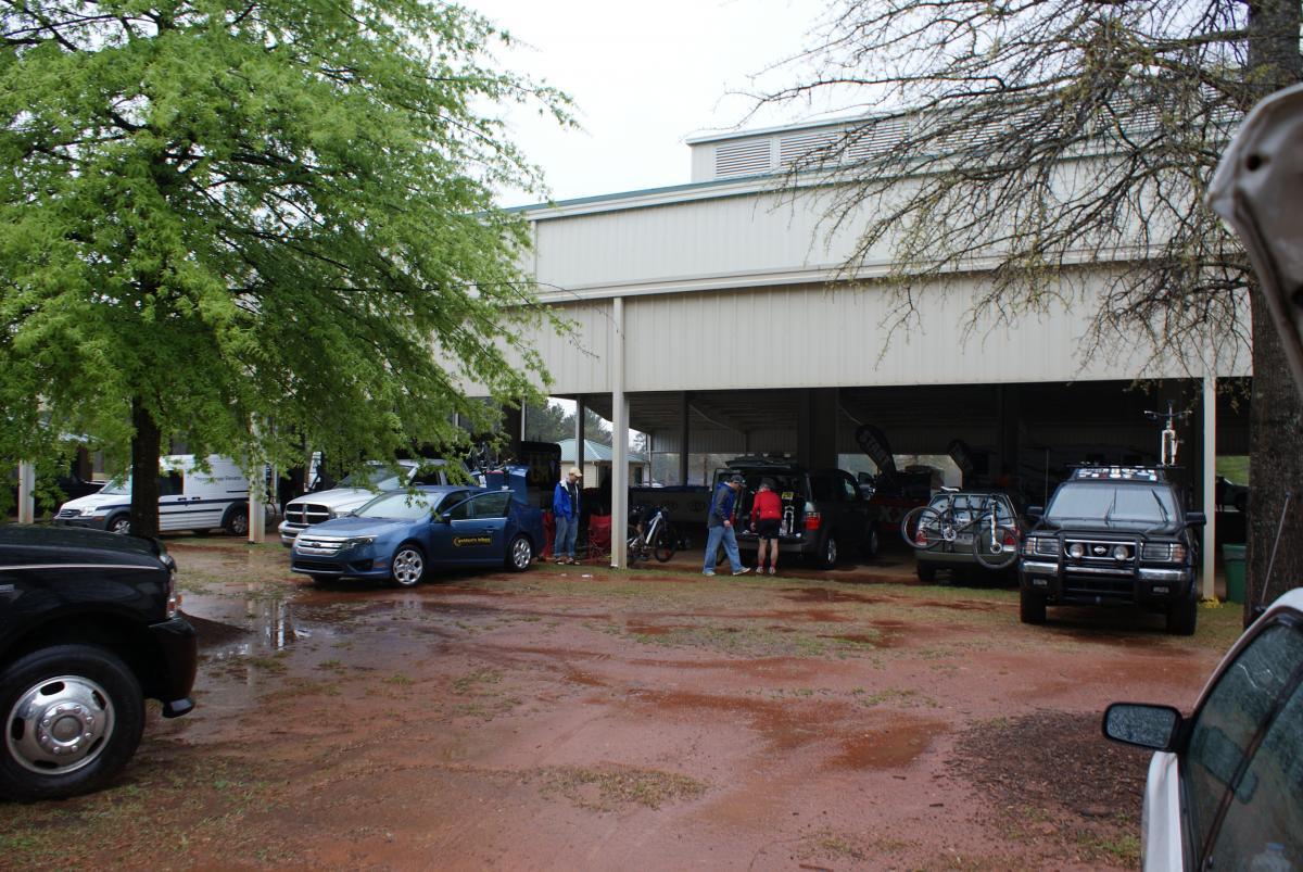 A parking area with several vehicles, including cars and SUVs, under a covered structure. People are gathered near the vehicles, some interacting and others preparing for an event. The ground is wet, suggesting recent rain, and there are green trees in the vicinity. Heritage Park mountain bike trail.