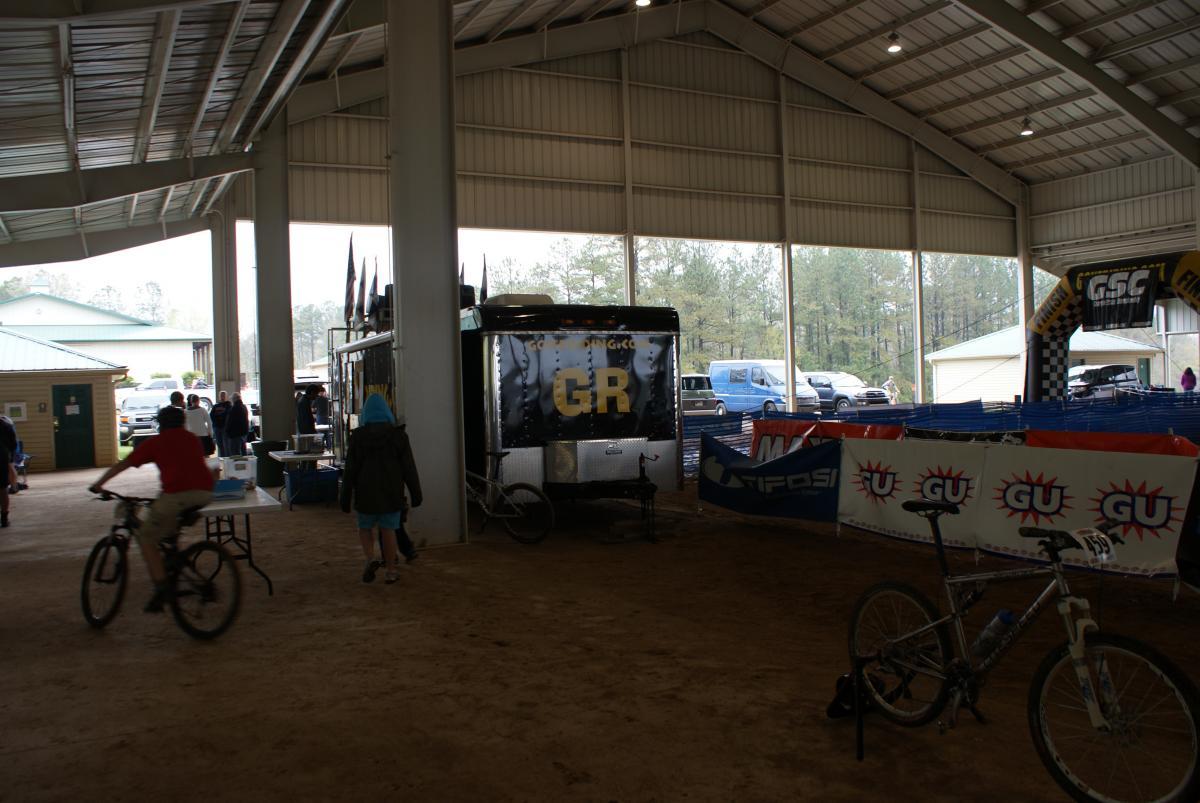 A large indoor space with a high ceiling, featuring various vendors and banners set up for an event. A person on a bicycle is riding past tables, while others are gathered nearby. At the back, a trailer with the letters "GR" is visible, along with parked vehicles and trees in the background. Heritage Park mountain bike trail.
