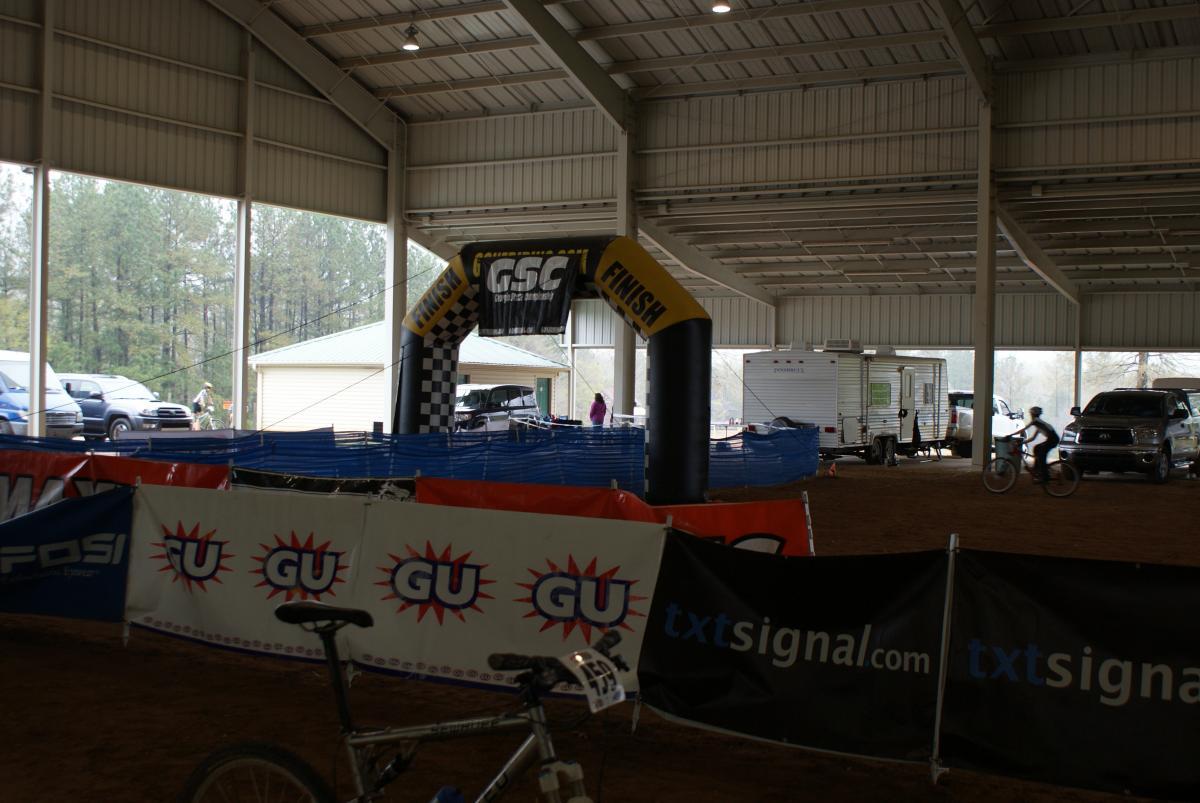 An indoor venue featuring a large inflatable finish arch marked with "FINISH" and "GSC," surrounded by vibrant event banners. In the foreground, a bicycle is visible, while a cyclist rides past in the background. The scene is set with vehicles parked nearby and trees visible through the open sides of the building. Heritage Park mountain bike trail.