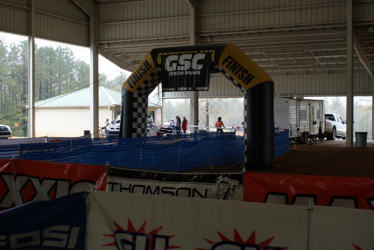 Inflatable finish line archway at a sporting event under an indoor structure, with various banners and a blue safety barrier in the foreground. Participants and spectators can be seen in the background, along with vehicles and a camper. Heritage Park mountain bike trail.