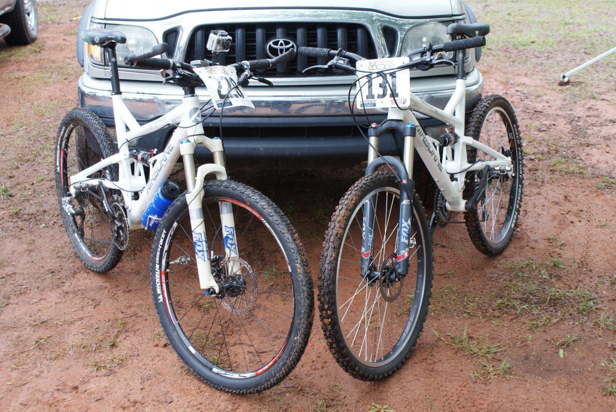 Two mountain bikes are parked side by side on a dirt surface, with a silver Toyota vehicle visible in the background. The bikes have white frames with distinct tire patterns and are equipped with front suspension forks. Each bike has a race number attached to the handlebars, indicating they may be used in a cycling event. The ground appears damp, suggesting recent rainfall. Heritage Park mountain bike trail.