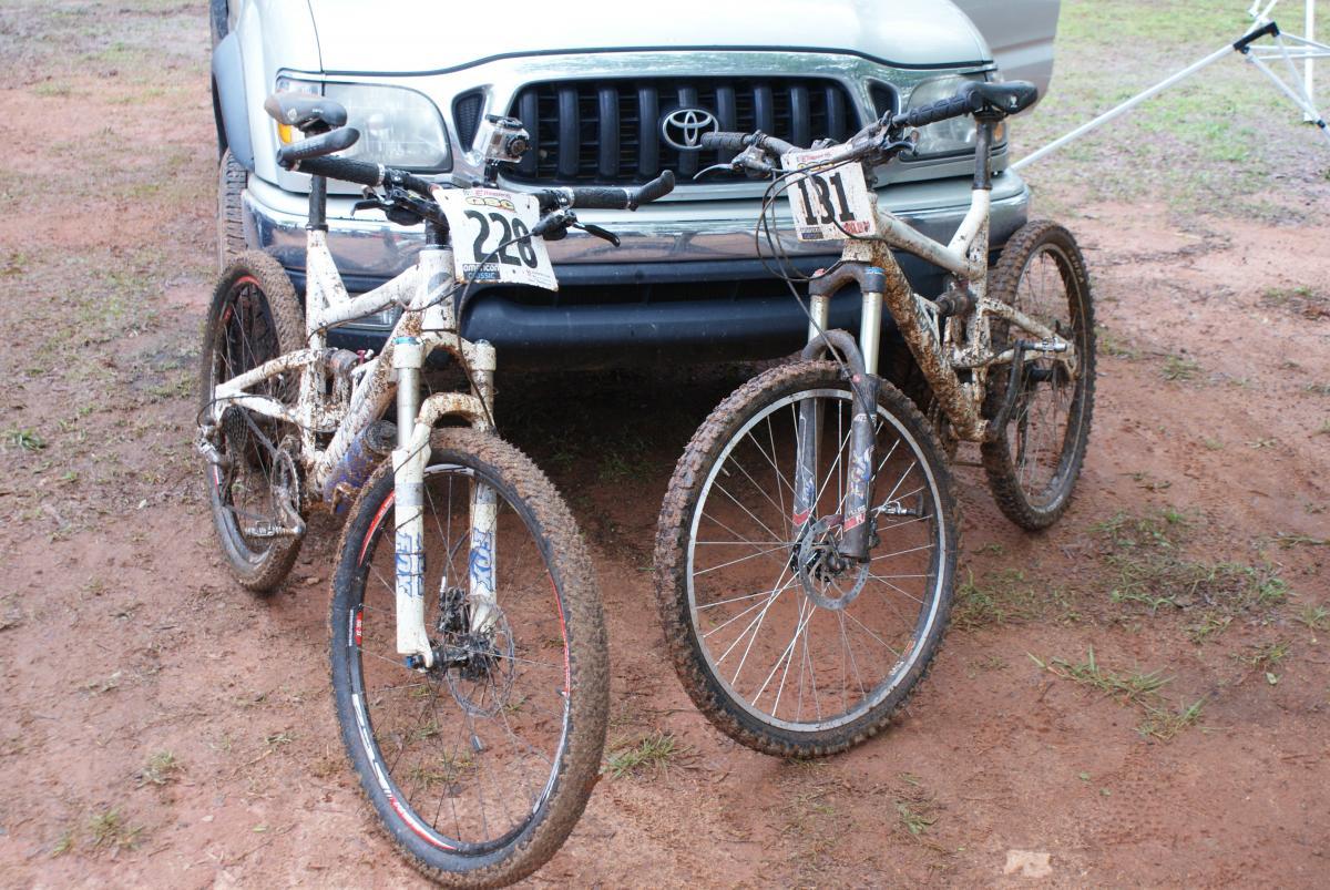 Two muddy mountain bikes with race numbers (228 and 111) parked in front of a silver Toyota truck. The bikes are resting on a dirt surface, showcasing signs of recent off-road use. Heritage Park mountain bike trail.