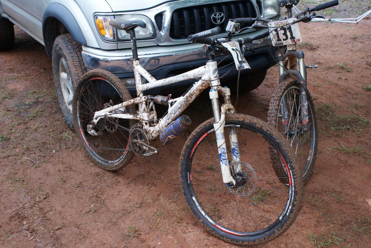 A muddy mountain bike parked next to a pickup truck. The bike has a white frame and is covered in dirt, with grass and mud on the ground beneath it. Another mountain bike is partially visible in the background, also showing signs of mud and use. Heritage Park mountain bike trail.