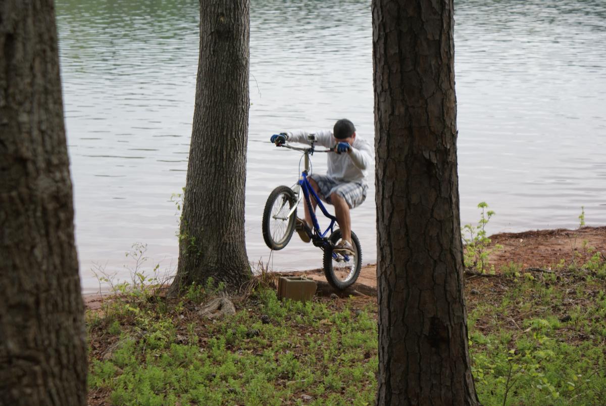 A young boy performing a wheelie on a blue bicycle near a calm lake, surrounded by trees and greenery. Lakeside Trail mountain bike trail.
