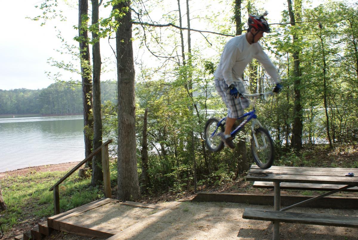 A person wearing a helmet and gloves performs a jump on a blue bicycle over a wooden picnic table on a gravel path by a lake, surrounded by trees and greenery. The scene captures an action shot during daytime, with a calm water background. Lakeside Trail mountain bike trail.