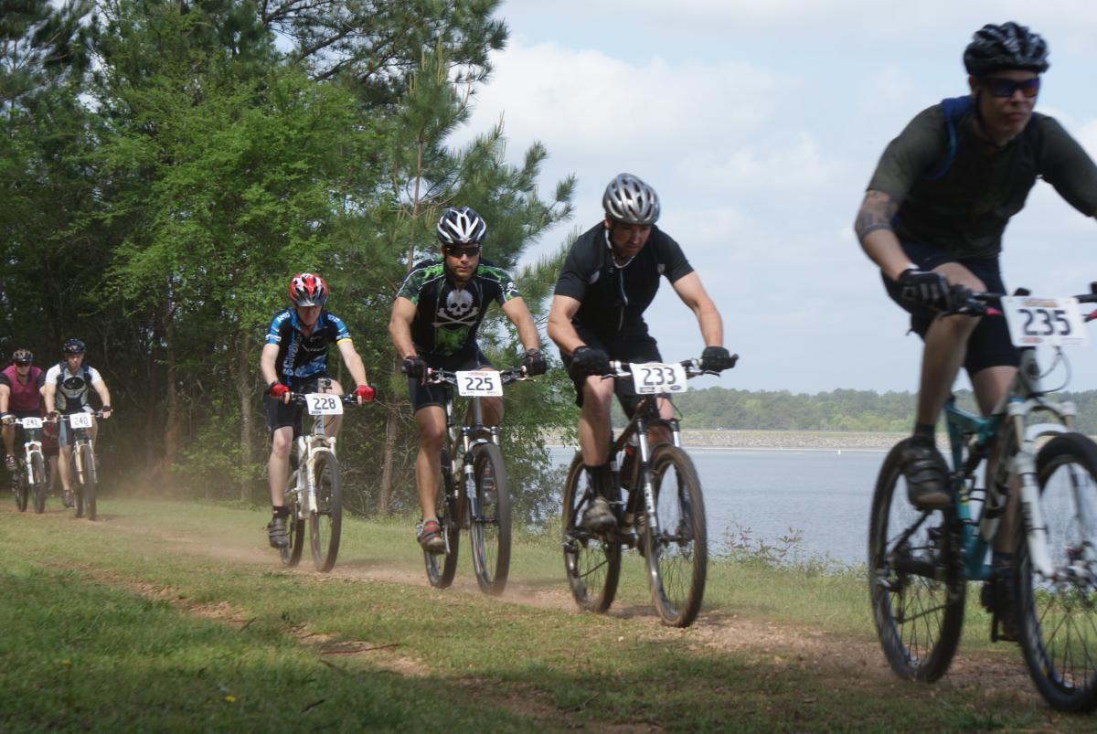 A group of six mountain bikers racing along a dirt trail beside a lake, surrounded by greenery. They are wearing helmets and cycling gear, with numbered bibs visible on their jerseys. Dust is kicked up from the trail as they pedal forward under a partly cloudy sky. Lakeside Trail mountain bike trail.