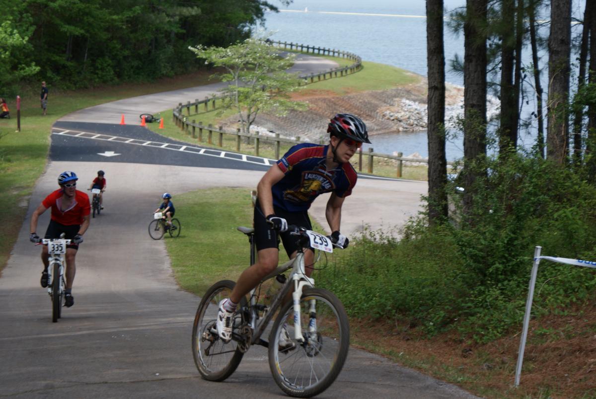A group of mountain bikers riding on a winding path through a forested area, with some riders climbing uphill and others pedaling in the background. A serene lake is visible nearby, and the scene is filled with greenery and trees. The cyclists are wearing helmets and jerseys, indicating a competitive event. Lakeside Trail mountain bike trail.