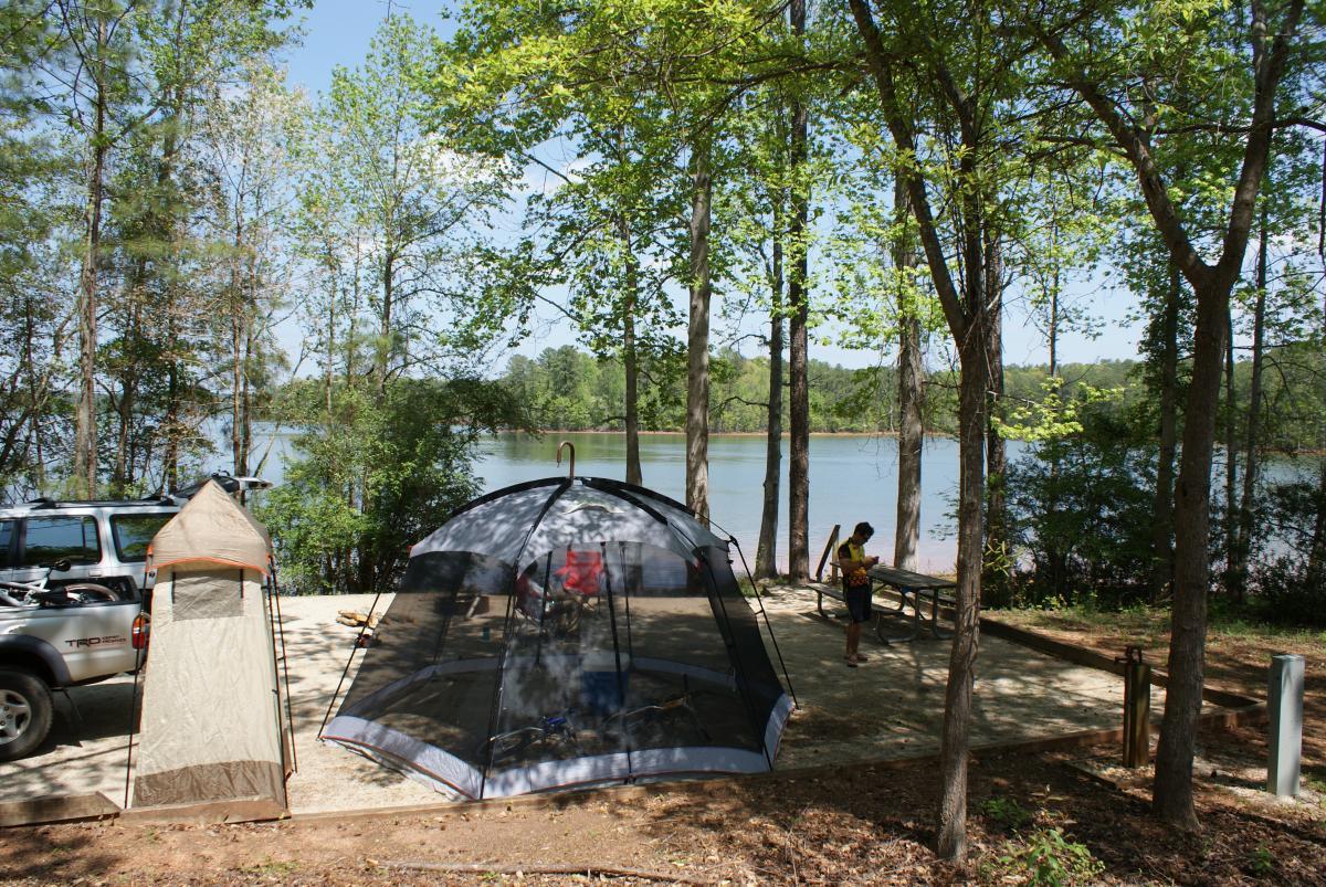 A peaceful lakeside campsite featuring a large tent with a mesh design, a smaller tent, and a parked vehicle. Lush green trees surround the area, and a lake is visible in the background under a clear blue sky. A person stands nearby, engaging with their surroundings. Lakeside Trail mountain bike trail.