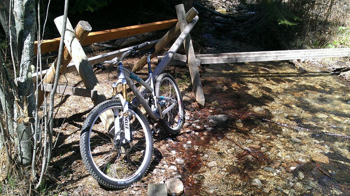 Yeti SB95: A mountain bike leaning against a wooden fence near a shallow, clear creek surrounded by trees and natural foliage. The sun is shining, creating a bright and inviting outdoor scene.