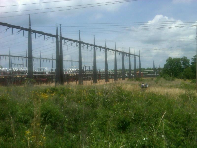 Two people walking through a grassy area in the foreground, with a large industrial structure featuring tall supports and power lines in the background under a partly cloudy sky. Raccoon Mountain Trail Network mountain bike trail.
