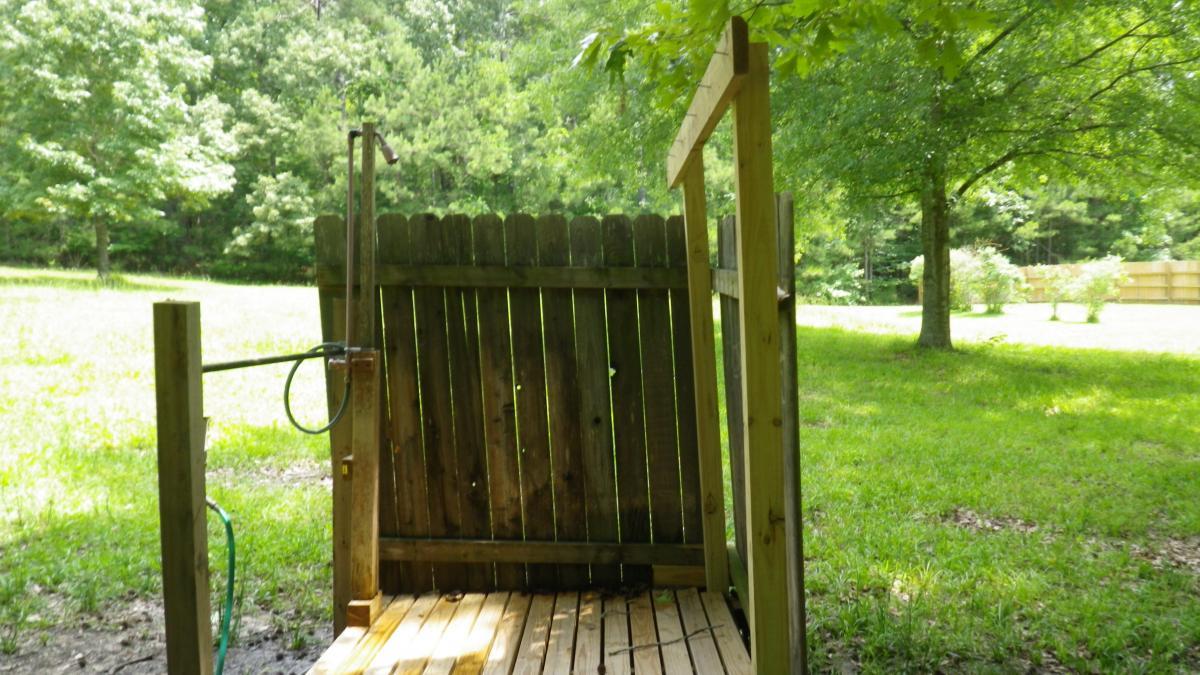 A wooden outdoor shower structure enclosed by a fence, with green grass and trees in the background. The shower features a simple wooden floor and a showerhead attached to a post. Natural light filters through the surrounding foliage, creating a serene and private setting. The Ridgeland Trails mountain bike trail.