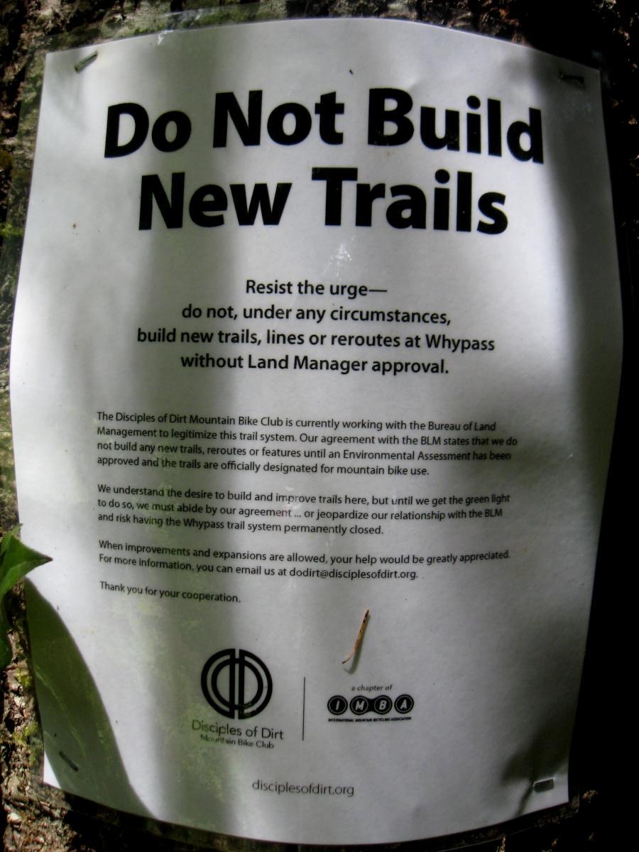A sign attached to a tree that reads "Do Not Build New Trails." The sign emphasizes the importance of not constructing new trails or reroutes without approval from the Land Manager. It mentions the Disciples of Dirt Mountain Bike Club's agreement with the Bureau of Land Management regarding trail management and encourages cooperation from users. The sign includes contact information for further inquiries. Whypass mountain bike trail.