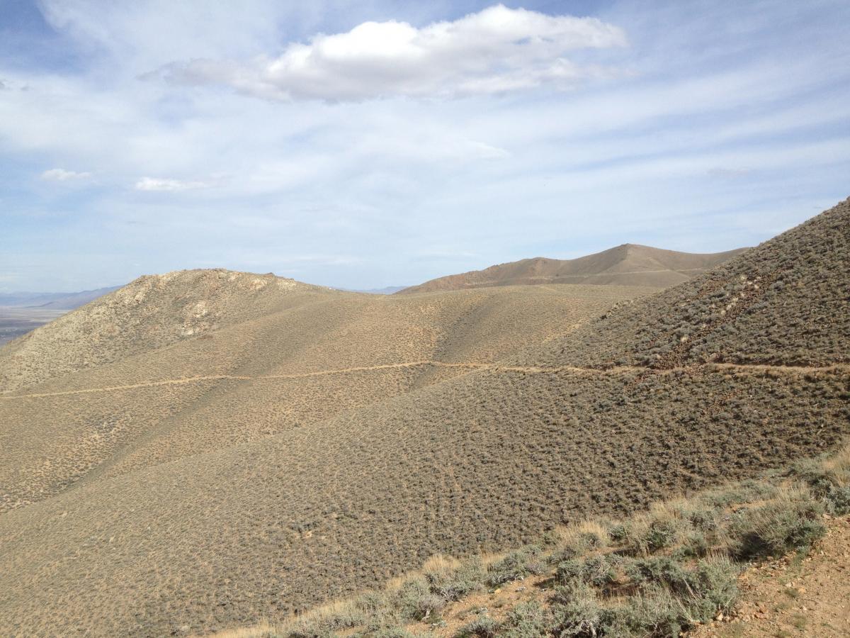 A panoramic view of rolling hills and mountains under a partly cloudy sky. The landscape features dry, sandy terrain with sparse vegetation and a winding dirt path visible along the slopes, showcasing the natural beauty of a remote area. Copper Basin Trail 3: Poormans Gulch mountain bike trail.