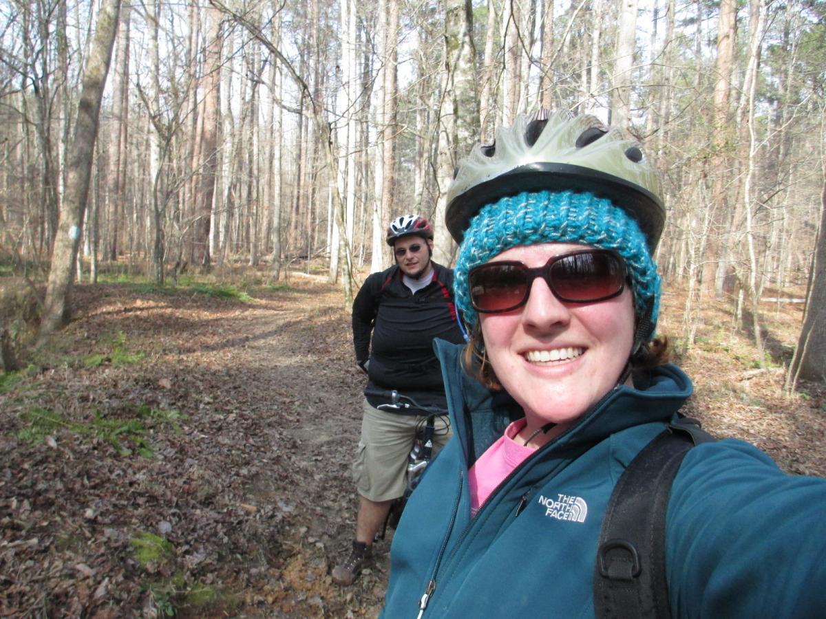 A woman wearing a helmet and sunglasses smiles at the camera while taking a selfie on a biking trail in a wooded area. In the background, a man dressed in a black sweater and cycling helmet stands next to a bike, with trees and fallen leaves surrounding them on the path. Buncombe - Brickhouse Recreation Area mountain bike trail.
