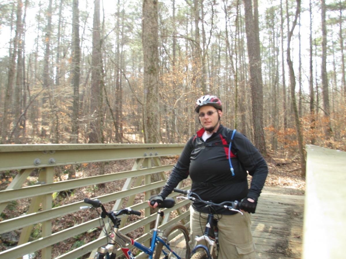 A person wearing a helmet and cycling gear stands on a bridge in a wooded area, holding a mountain bike. The bridge is made of wood and overlooks a small creek, with tall trees and autumn foliage in the background. Buncombe - Brickhouse Recreation Area mountain bike trail.