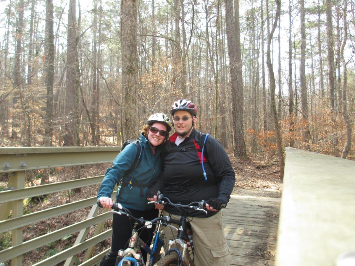 A couple smiles while standing on a wooden bridge in a forested area, each with a mountain bike beside them. They are wearing cycling helmets and casual outdoor clothing, surrounded by tall trees and fall foliage. The atmosphere is cheerful and adventurous. Buncombe - Brickhouse Recreation Area mountain bike trail.
