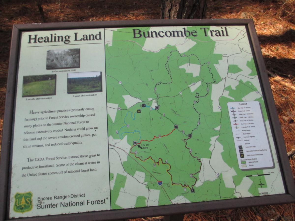 Alt text: A sign titled "Healing Land Buncombe Trail" displaying information about land restoration in the Sumter National Forest. The sign includes before-and-after images of the land, a brief explanation of agricultural practices that caused erosion, and details on the USDA Forest Service's efforts in restoring the area. A map shows the trail layout and surrounding features. Buncombe - Brickhouse Recreation Area mountain bike trail.