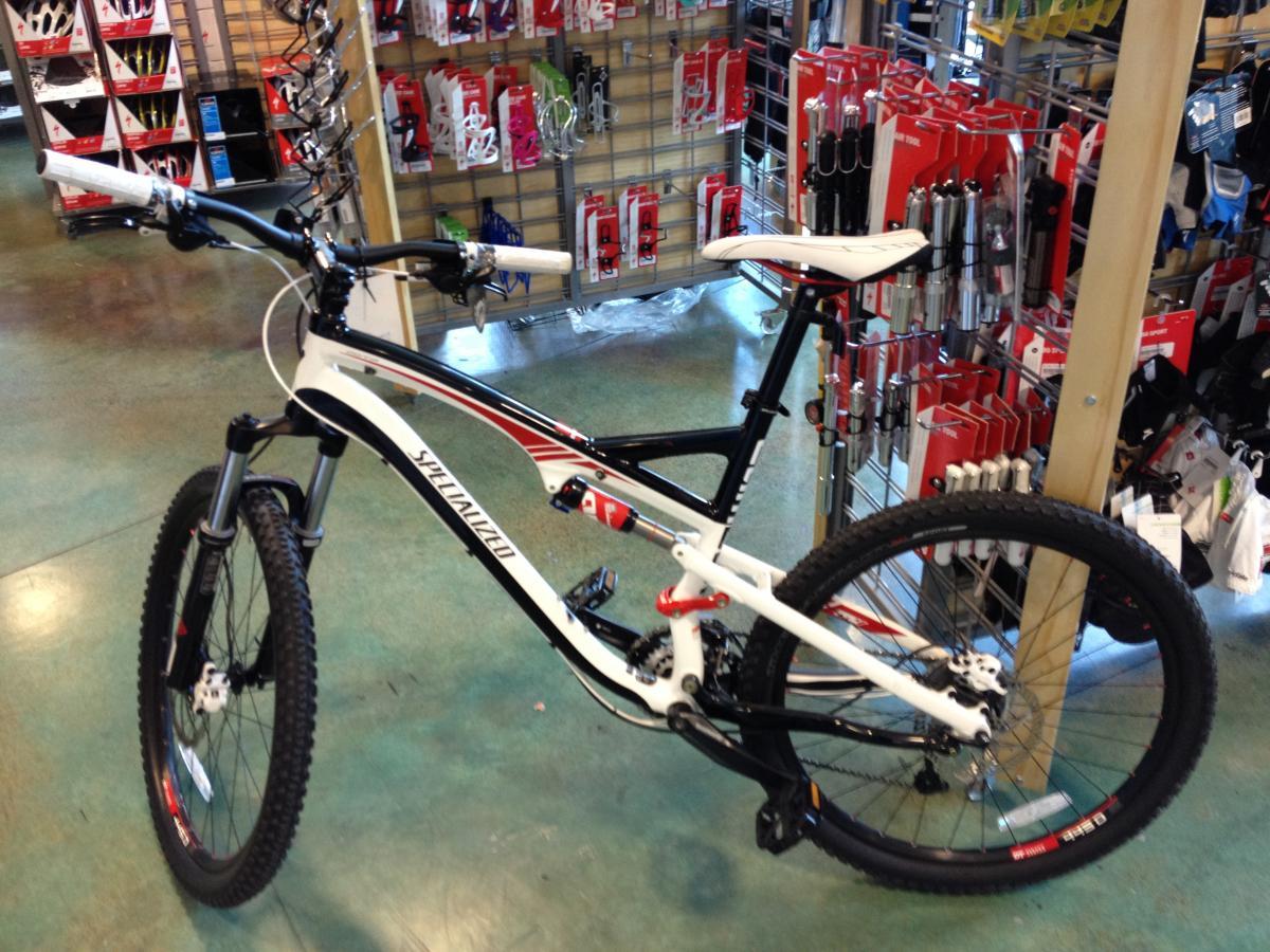 Specialized Camber: A mountain bike displayed in a retail store, featuring a sleek design with a white and black frame and red accents. The bike has thick tires for off-road capability, suspension forks, and is positioned next to a display of cycling accessories. The store background contains various biking gear and tools.