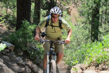 A person in a helmet rides a mountain bike along a rocky trail surrounded by trees and greenery. They appear focused as they navigate the challenging terrain. Colorado Trail: Kennebec Pass To Junction Creek mountain bike trail.