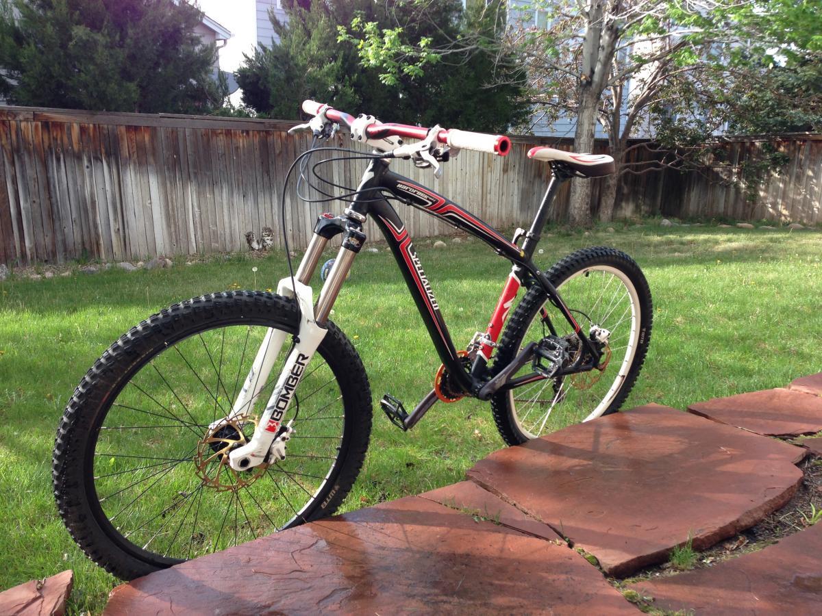 Specialized Hardrock Disc: A mountain bike with a red and black frame is standing on a grassy area next to stone pavers. The bike features thick, knobby tires and front suspension components. In the background, there is a wooden fence and some shrubs.