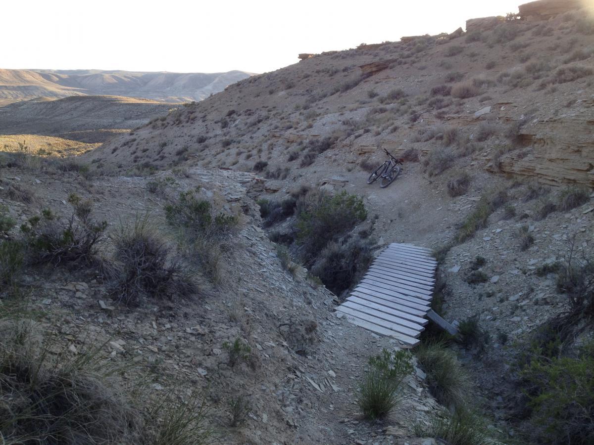 A dirt trail winding through a rugged landscape, featuring a wooden bridge crossing a shallow ravine. In the background, rolling hills and vegetation are visible, along with a mountain bike leaning against a rocky outcrop. The scene is illuminated by soft sunlight, creating a serene outdoor atmosphere. Brent & Mikes' Trail mountain bike trail.