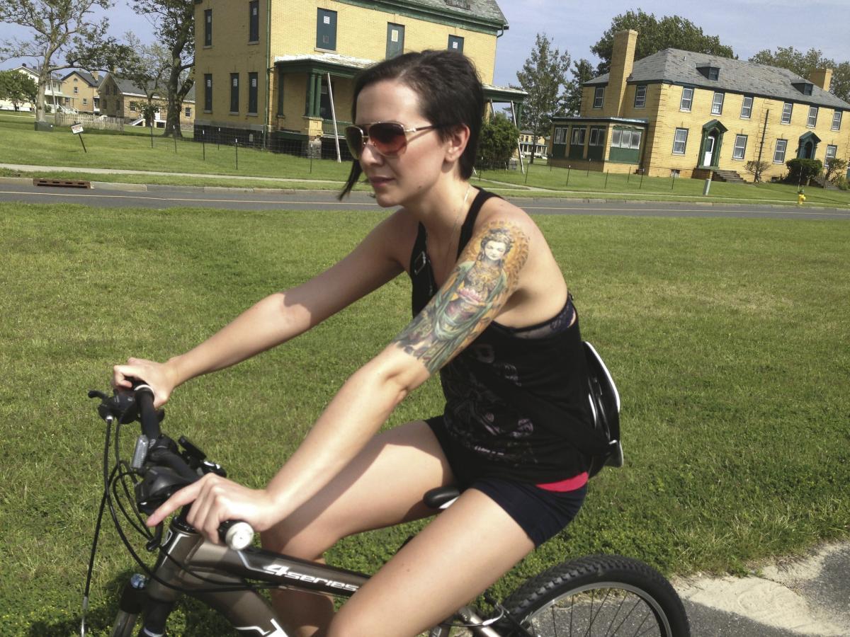 A young woman with short hair and sunglasses is riding a mountain bike on a grassy area. She has a colorful tattoo on her left arm and is wearing a black tank top and shorts. In the background, there are yellow buildings and trees under a clear sky.