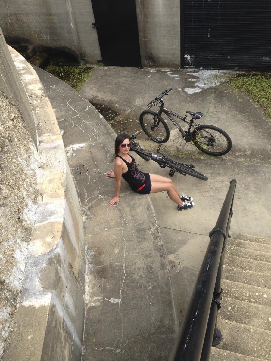 A person sitting on a concrete surface with a bike nearby, wearing sunglasses and casual athletic clothing. The background features a set of stairs and a black door, with patches of greenery visible on the ground.