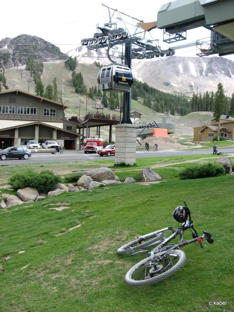 A mountain bike rests on green grass in the foreground, with a ski lift visible in the background against a mountainous landscape. Buildings are present nearby, and a road with parked cars runs alongside the image. The scene captures a recreational area, likely for both biking and skiing. Mammoth Mountain Resort mountain bike trail.