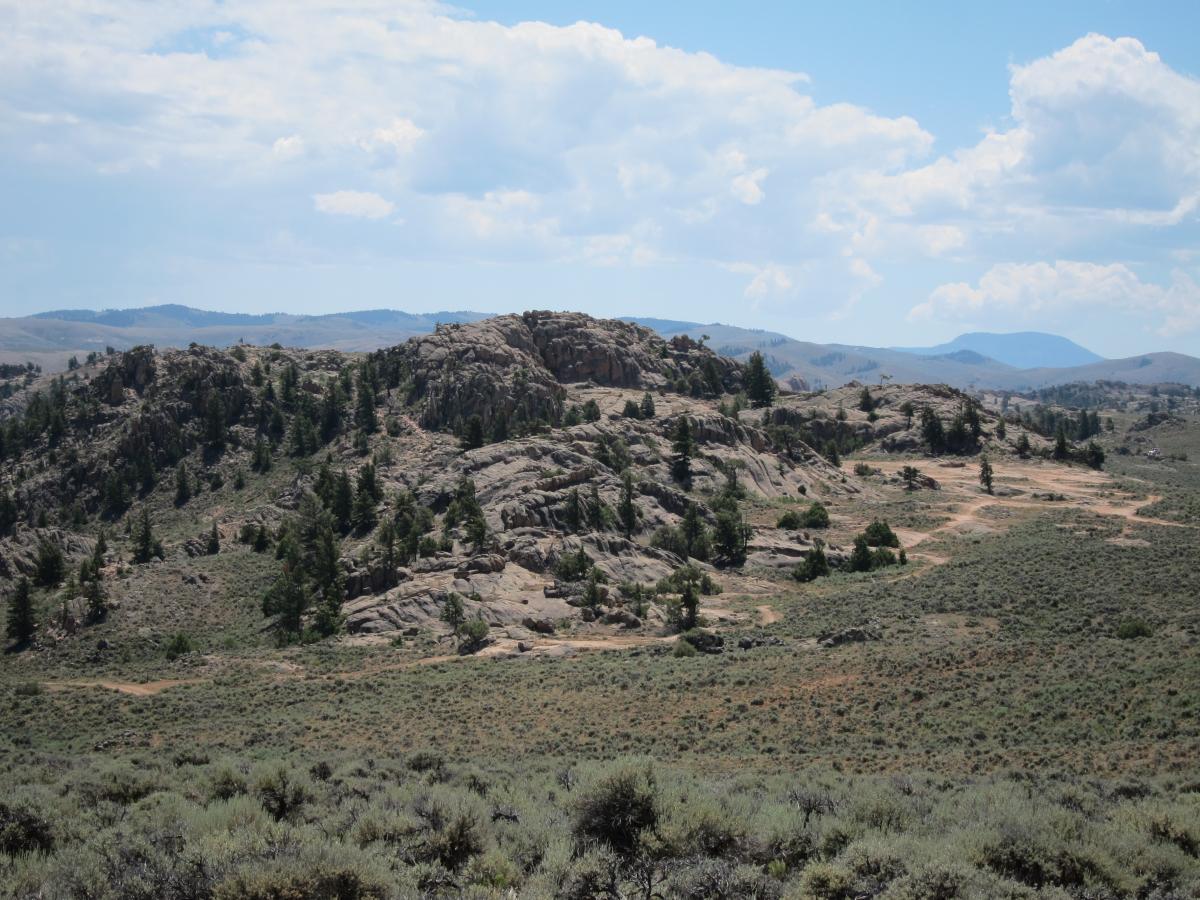 A mountainous landscape featuring rocky outcrops and sparse vegetation under a partly cloudy sky. The foreground shows rolling hills covered with low shrubs, while the background consists of distant mountain ranges. Hartman Rocks mountain bike trail.