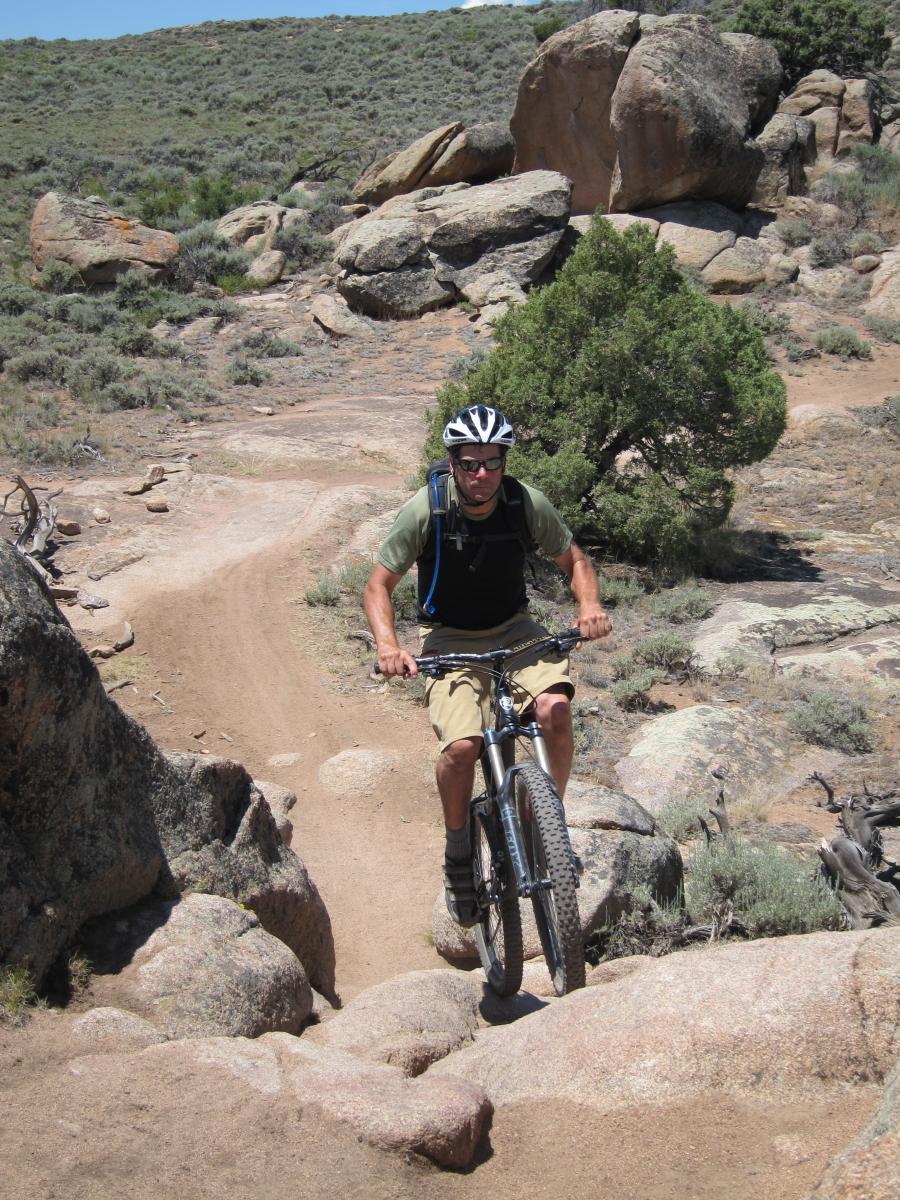 A mountain biker navigating a rocky trail in a mountainous landscape. The cyclist, wearing a helmet and sunglasses, is focused as he approaches a rocky incline surrounded by shrubs and boulders under a clear blue sky. Hartman Rocks mountain bike trail.