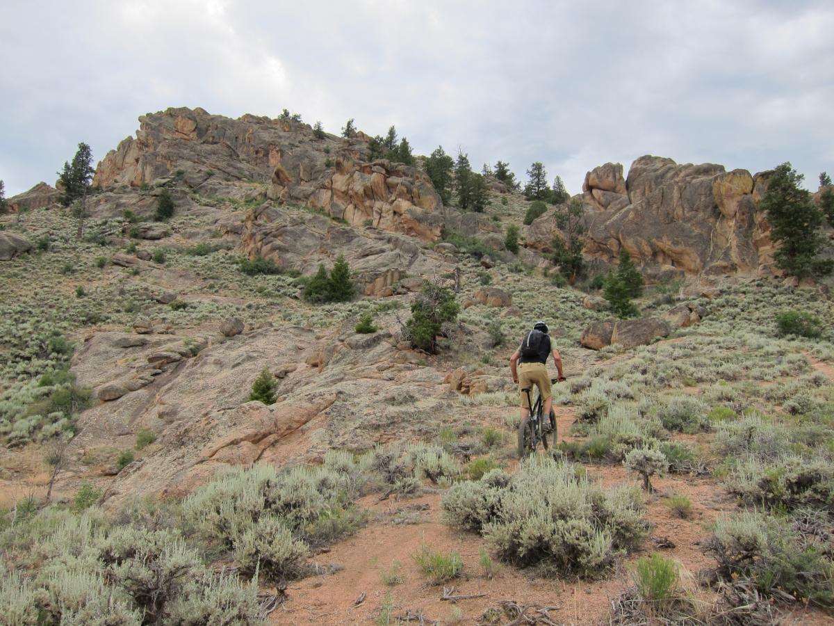 A person riding a mountain bike on a rocky trail surrounded by green shrubs and rocky terrain, with steep cliffs and trees in the background under a cloudy sky. Hartman Rocks mountain bike trail.