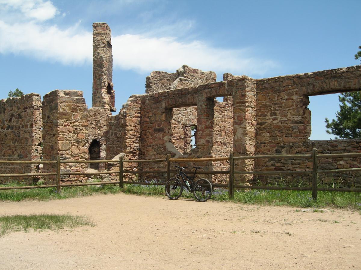 Alt text: A mountain bike parked in front of the ruins of a stone building, with a clear blue sky and wispy clouds in the background. A wooden fence surrounds the area, and greenery is visible in the foreground. Mount Falcon Park mountain bike trail.