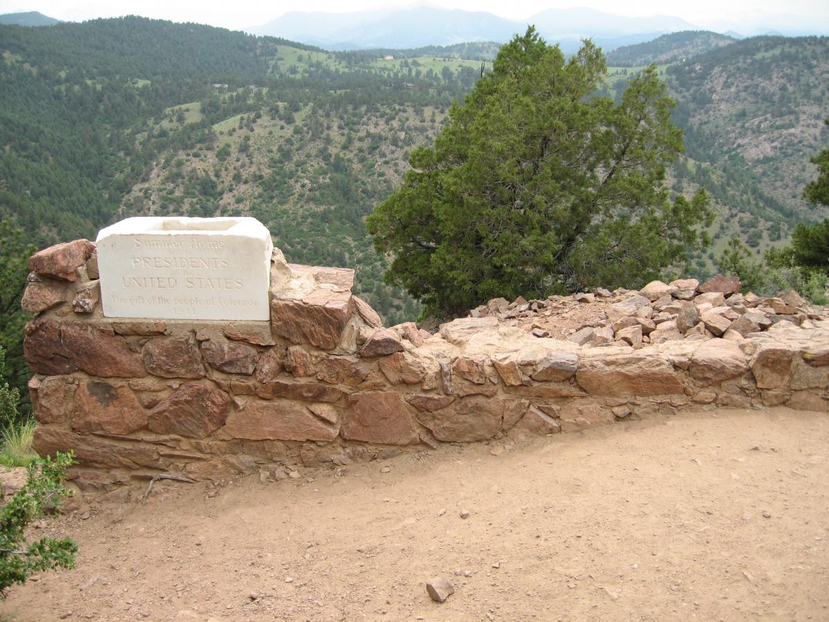 A stone wall with a plaque that reads "Summer Home Presidents United States The gift of the people of Colorado 1911." The background features lush green mountains and a cloudy sky, evoking a serene and historic setting. Mount Falcon Park mountain bike trail.