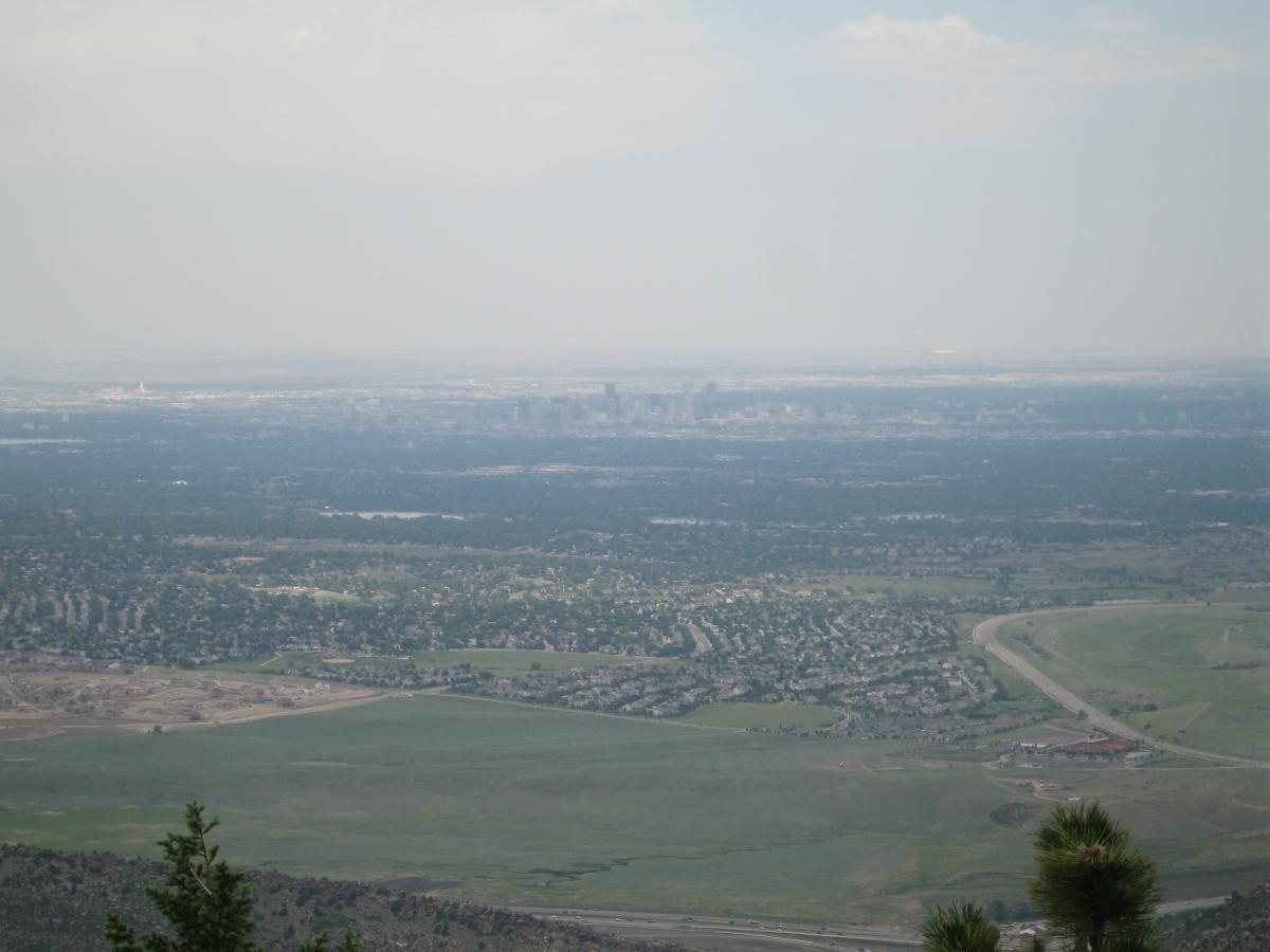 A panoramic view of a city skyline in the distance, surrounded by green fields and suburban neighborhoods. The city appears hazy and is situated against a backdrop of light clouds in the sky. In the foreground, there are patches of grass and scattered trees, indicating a higher vantage point overlooking the landscape below. Mount Falcon Park mountain bike trail.