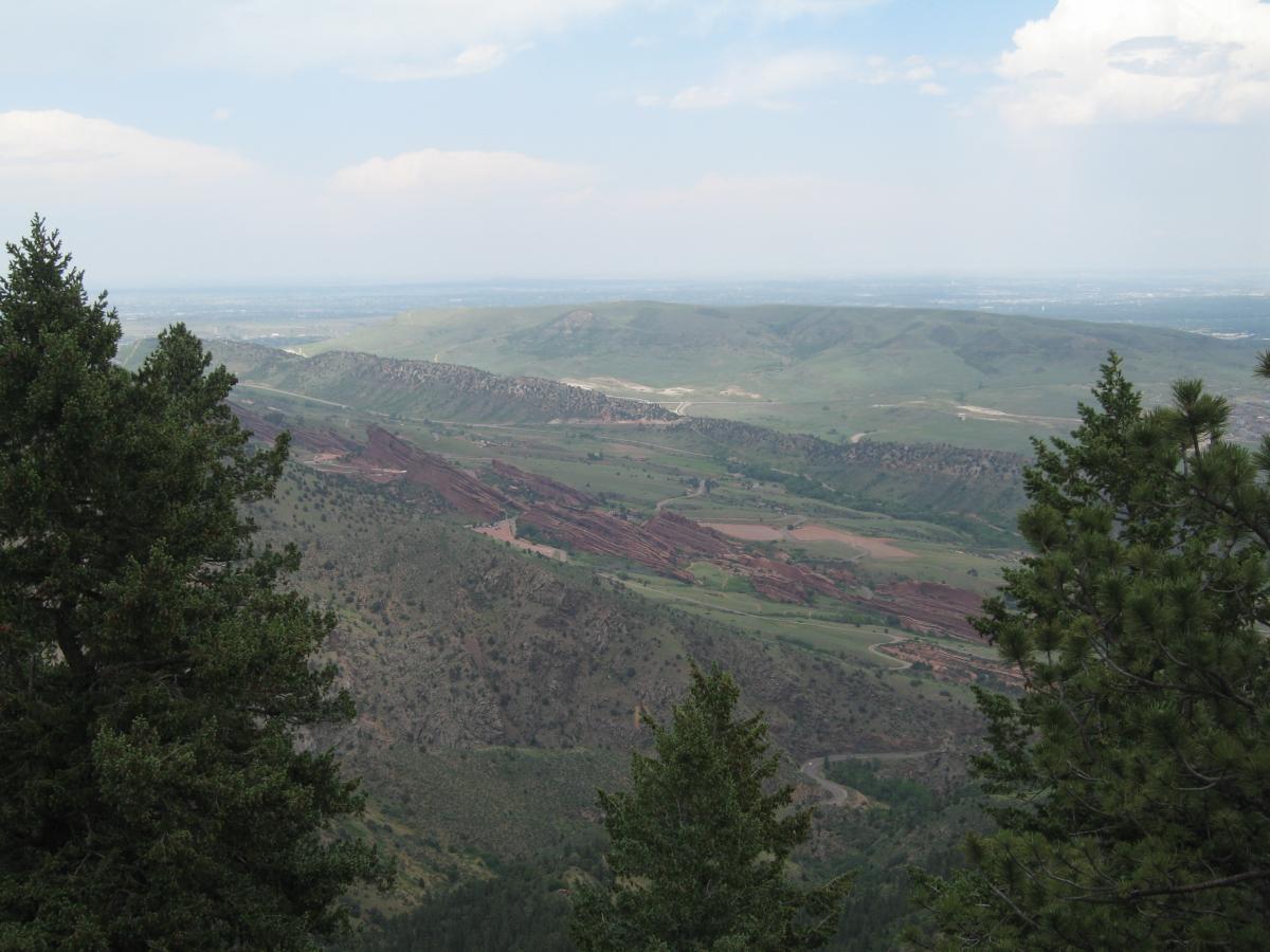 A panoramic view of rolling green hills and rocky landscapes under a partly cloudy sky, framed by evergreen trees on either side. The scene captures the natural beauty of a mountainous region with a visible winding road cutting through the terrain. Mount Falcon Park mountain bike trail.