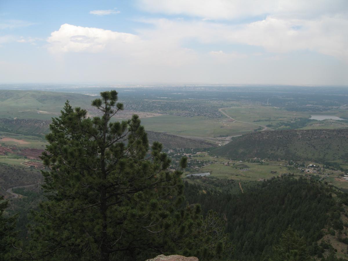 A panoramic view of a valley from a mountainous vantage point, featuring green hills and scattered residential areas in the foreground. The distant city skyline can be seen beneath a partly cloudy sky, with patches of blue peeking through the clouds. A conifer tree is prominently visible in the foreground, adding to the natural landscape. Mount Falcon Park mountain bike trail.