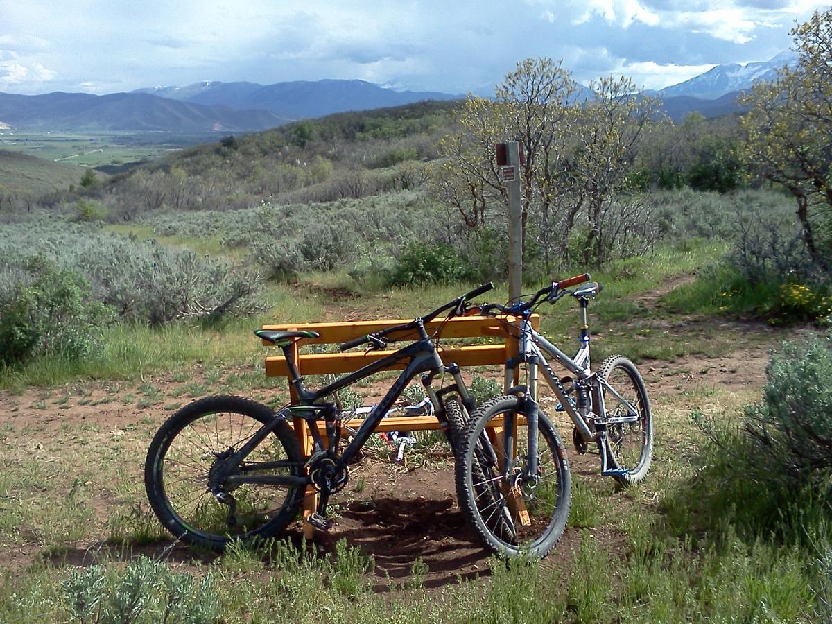 Trek Fuel EX 9.8: Two mountain bikes are parked against a wooden bench with scenic hills and mountains in the background. The landscape features green shrubs and trees under a cloudy sky, indicating an outdoor recreational area.