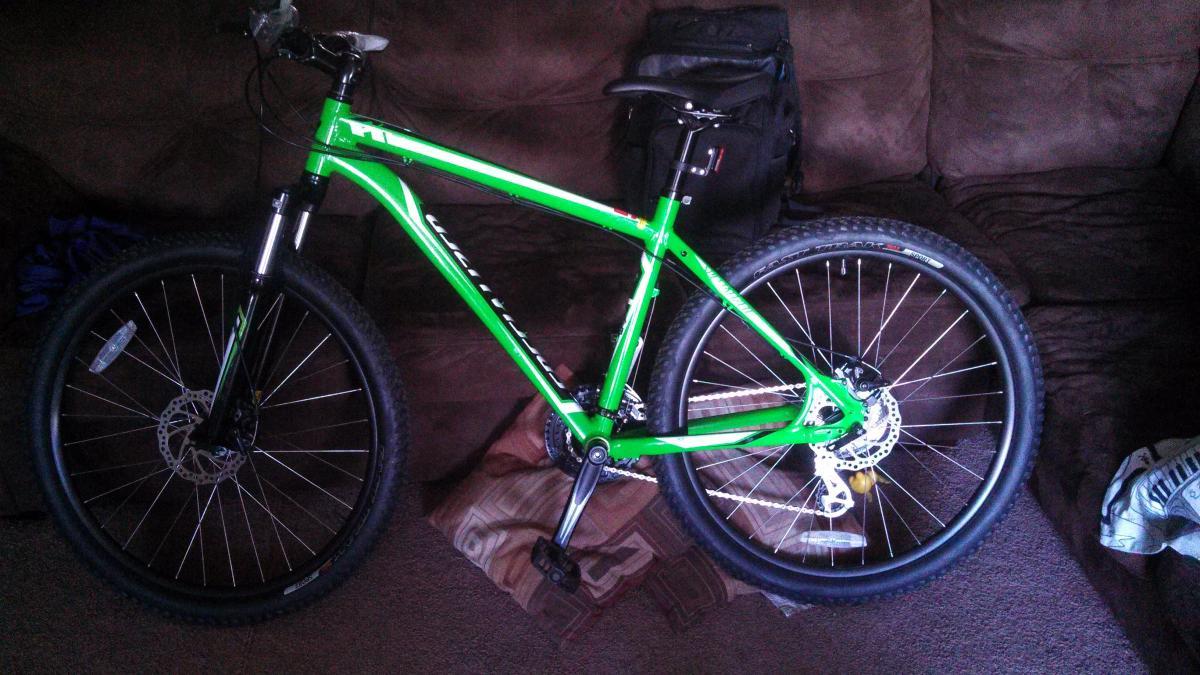 Specialized Hardrock Disc: A green mountain bike resting on a couch, with a black bag in the background. The bike features large tires, a lightweight frame, and disc brakes. The setting includes a casual living room environment with patterned cushions and a partially visible blanket.