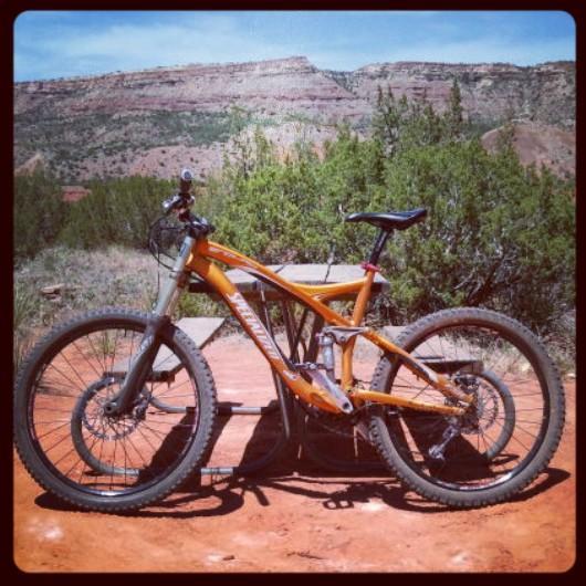 Specialized Enduro SL Pro: A bright orange mountain bike parked on a wooden ramp, with a scenic backdrop of red rock formations and greenery under a clear blue sky.
