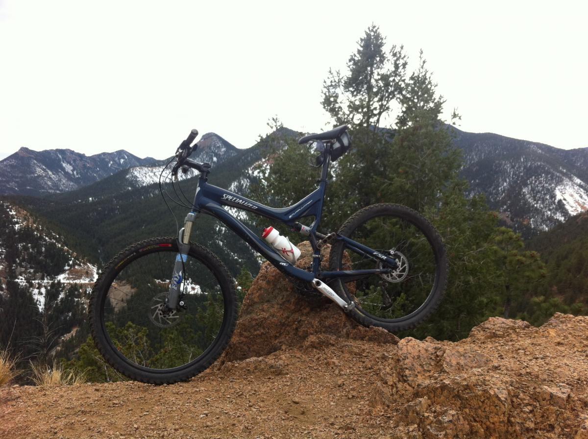 Specialized Stumpjumper FSR: A blue mountain bike is prominently positioned on a rocky outcrop, overlooking a mountainous landscape filled with pine trees and snow-capped peaks in the background. A water bottle is attached to the bike's frame, and the sky is overcast, suggesting a cool day for outdoor activities.